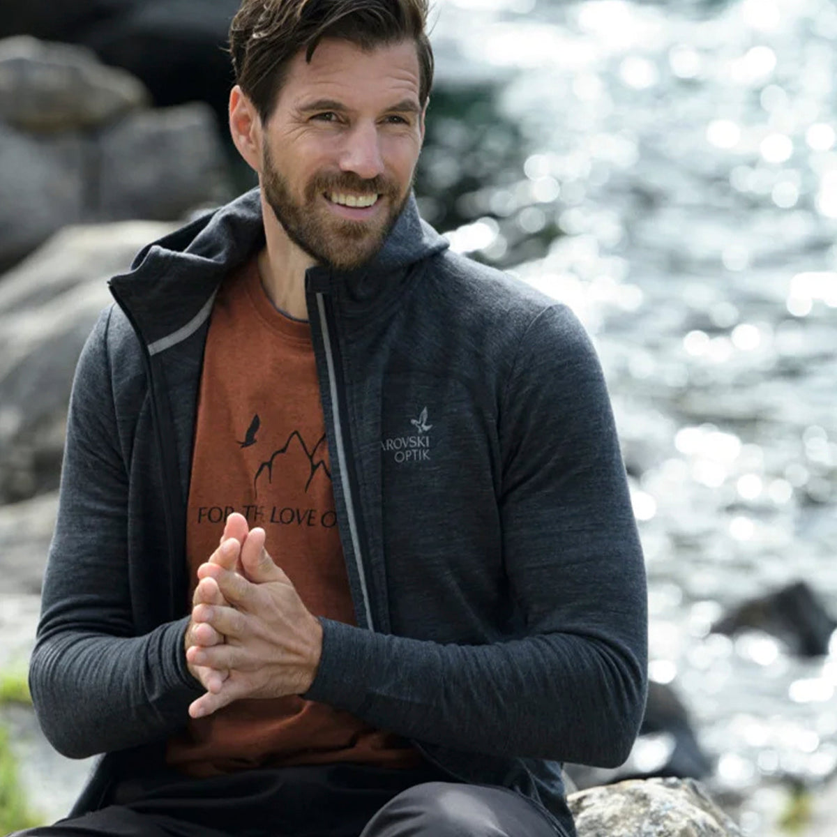 Hombre con sudadera Swarovski Optik MH Merino gris abierta, sentado junto al agua